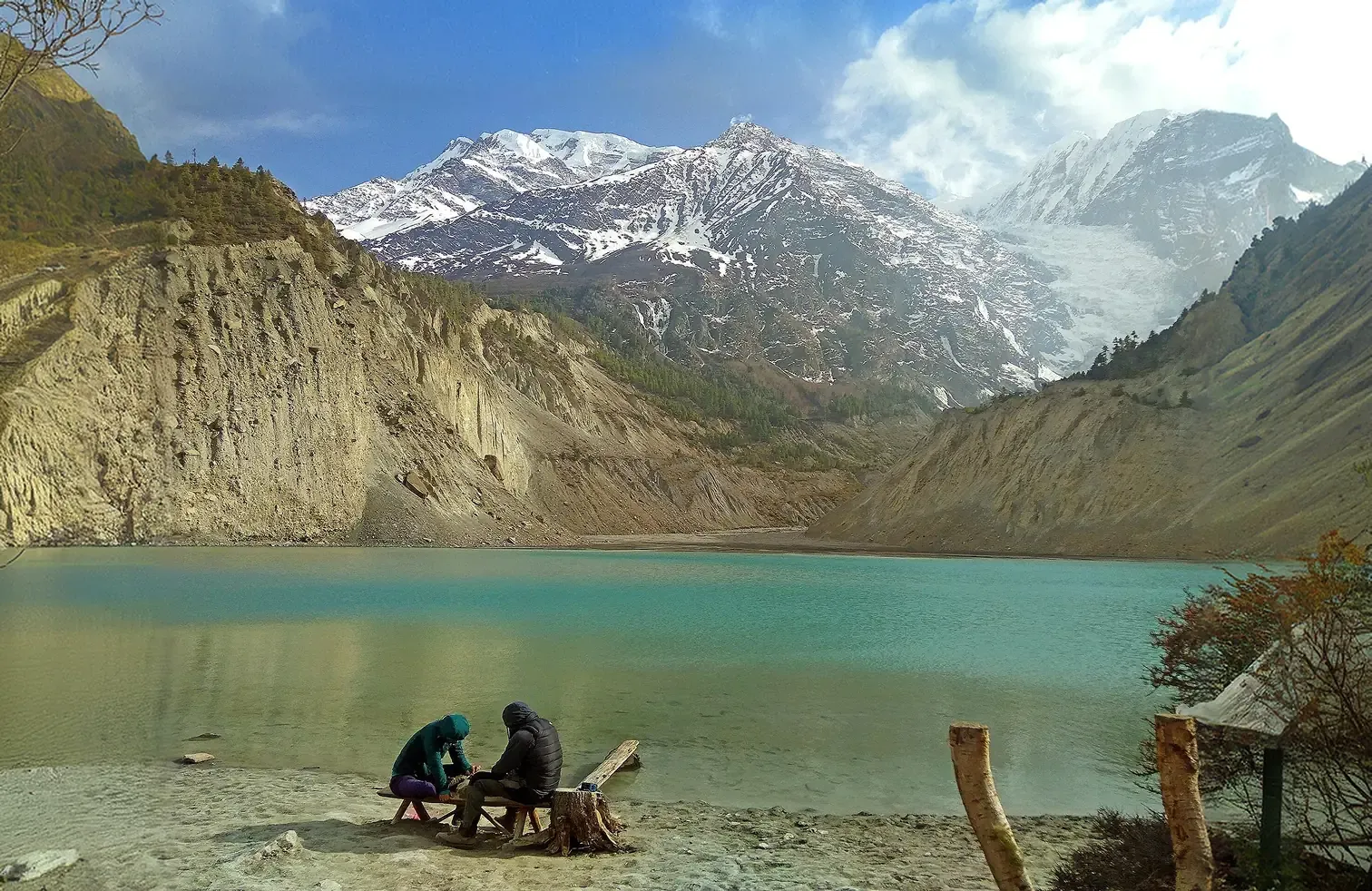 Turquoise glacial lake below the snow-capped Annapurna range, with two people seated at its shore in Manang